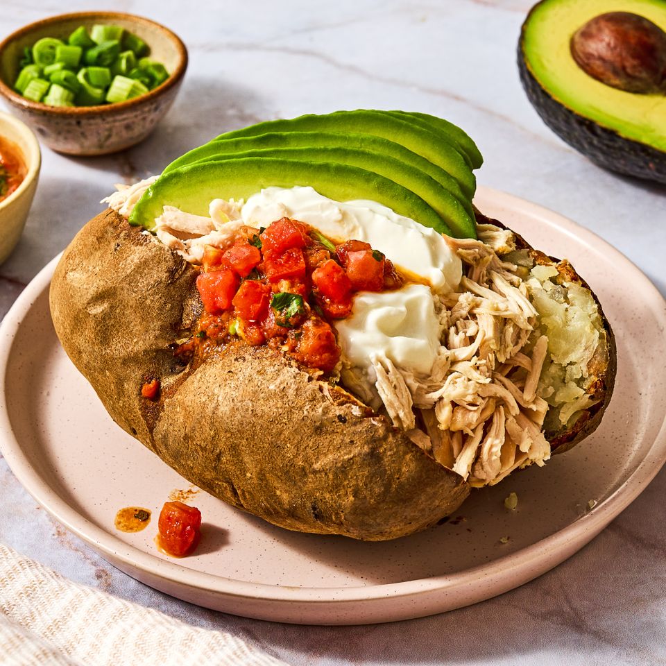 A stuffed baked potato topped with shredded chicken diced tomatoes sour cream and avocado slices with garnish bowls in the background