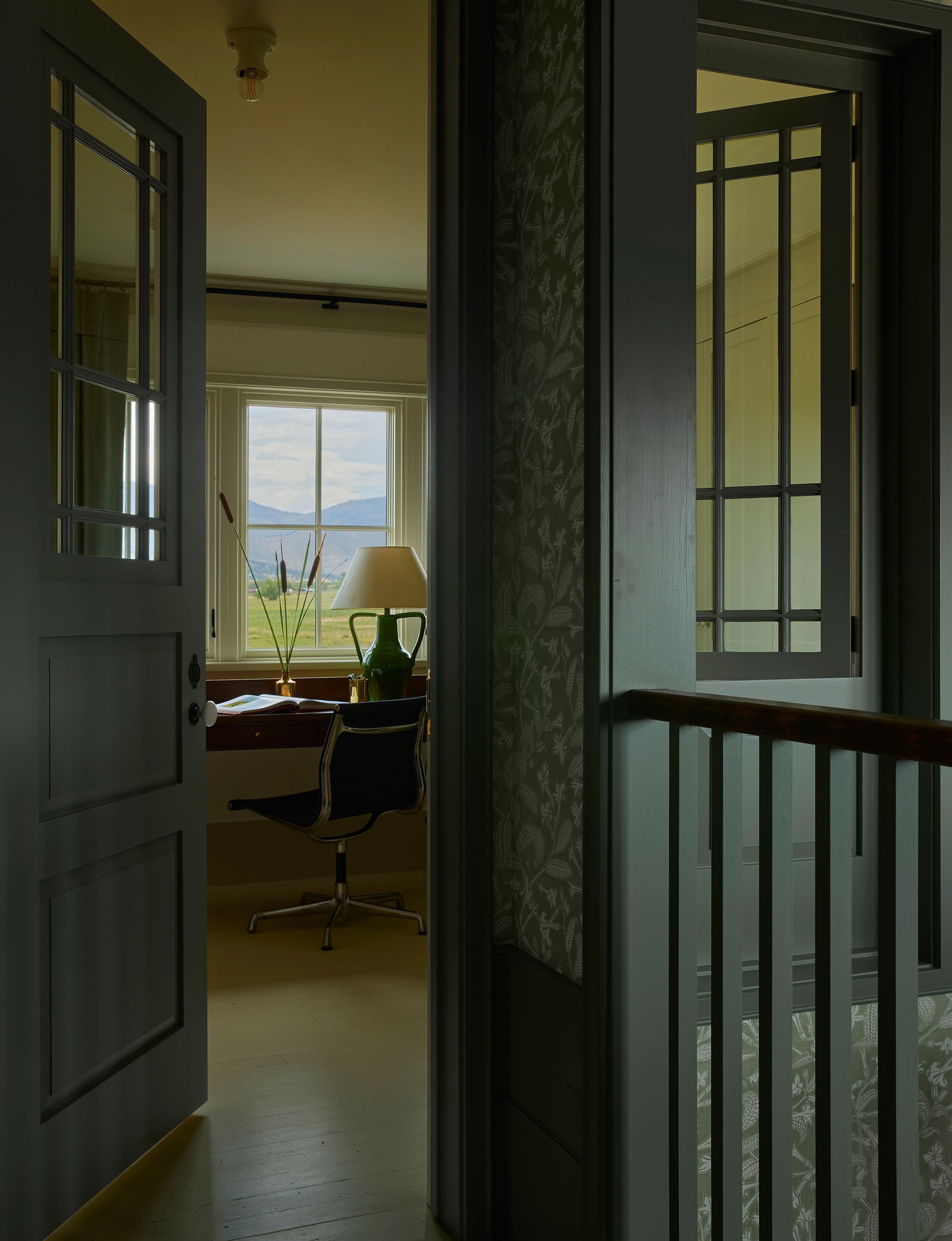 On the second-floor landing, an interior window implies a house with a smaller footprint, as if the house grew and changed over the years alongside a family. The trim and door is Card Room Green by Farrow & Ball. An iris lamp in fern by Nickey Kehoe sits on a custom desk by Landed Interiors made from a piece via Petersen Antiques.