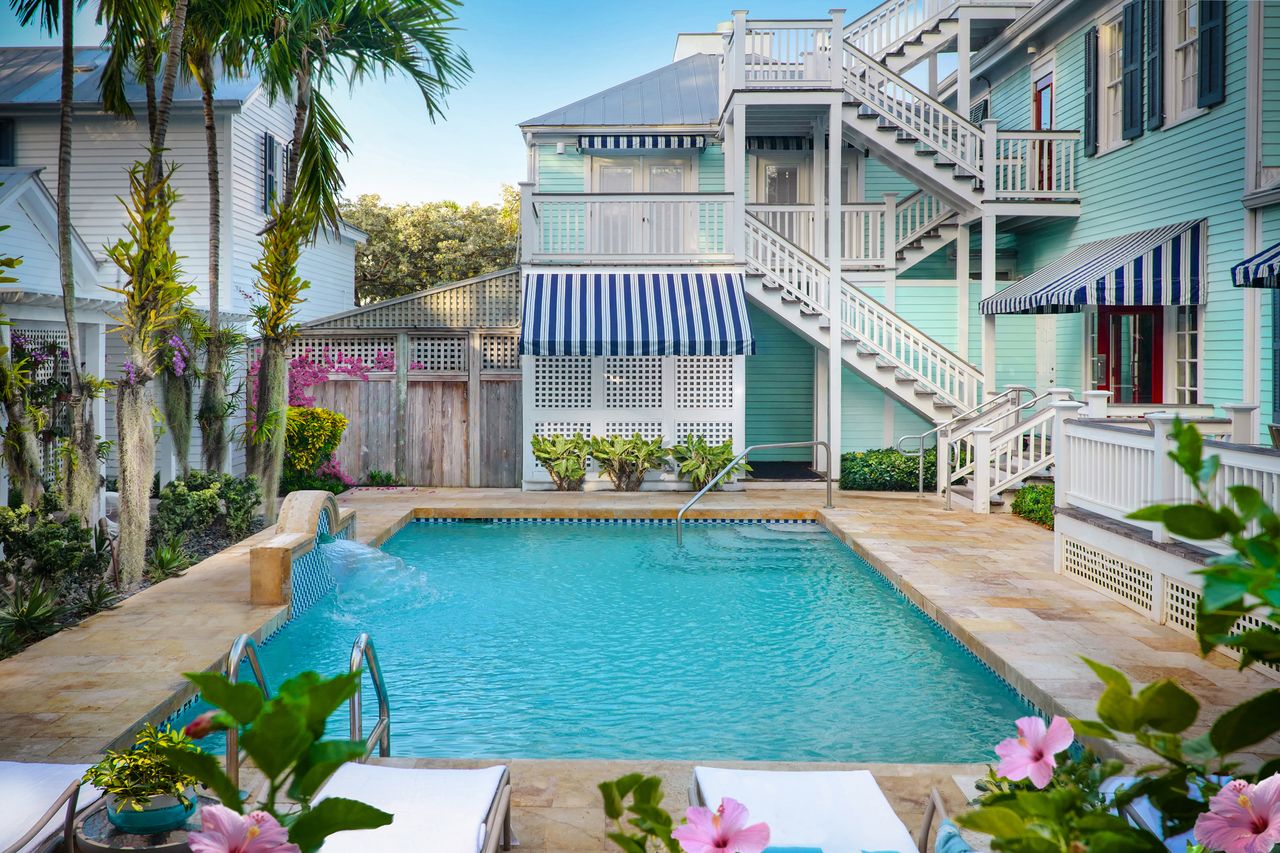 An outdoor pool surrounded by a residentialstyle building with striped awnings and a staircase tropical plants frame the scene