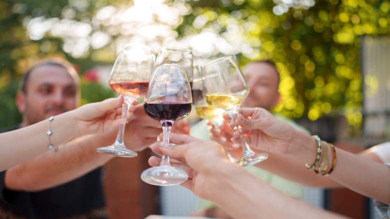 A close-up of six clinking wine glasses filled with different varietals, with out-of-focus people and sunlit green trees in the background.