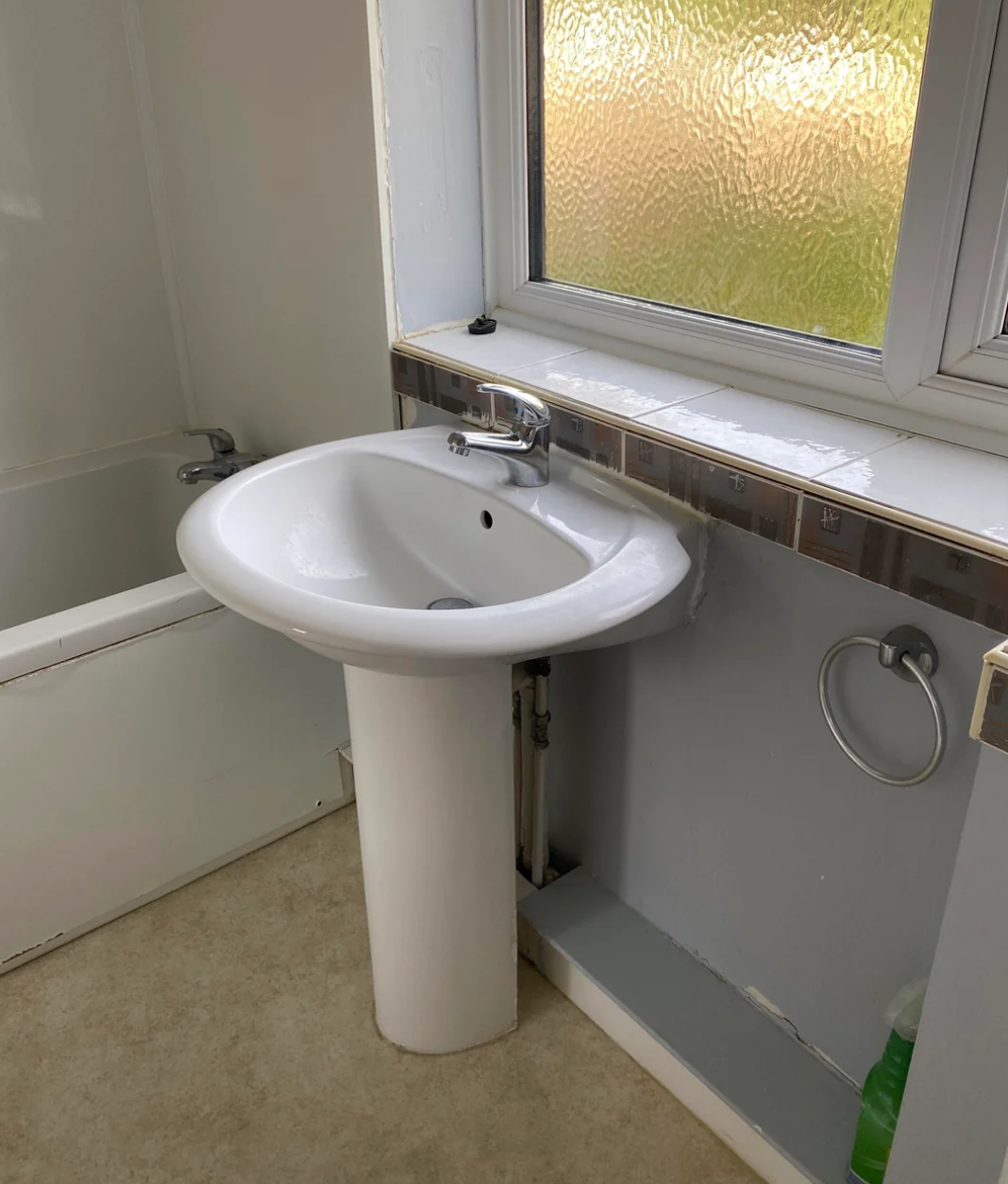 A bathroom with a pedestal sink, a frosted window, and a tub partially visible. There's a towel ring and a cleaning product by the sink