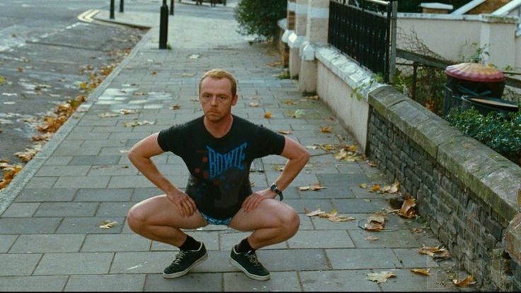 A man in a "Bowie" t-shirt humorously squats on an empty sidewalk, striking a playful pose in a casual setting