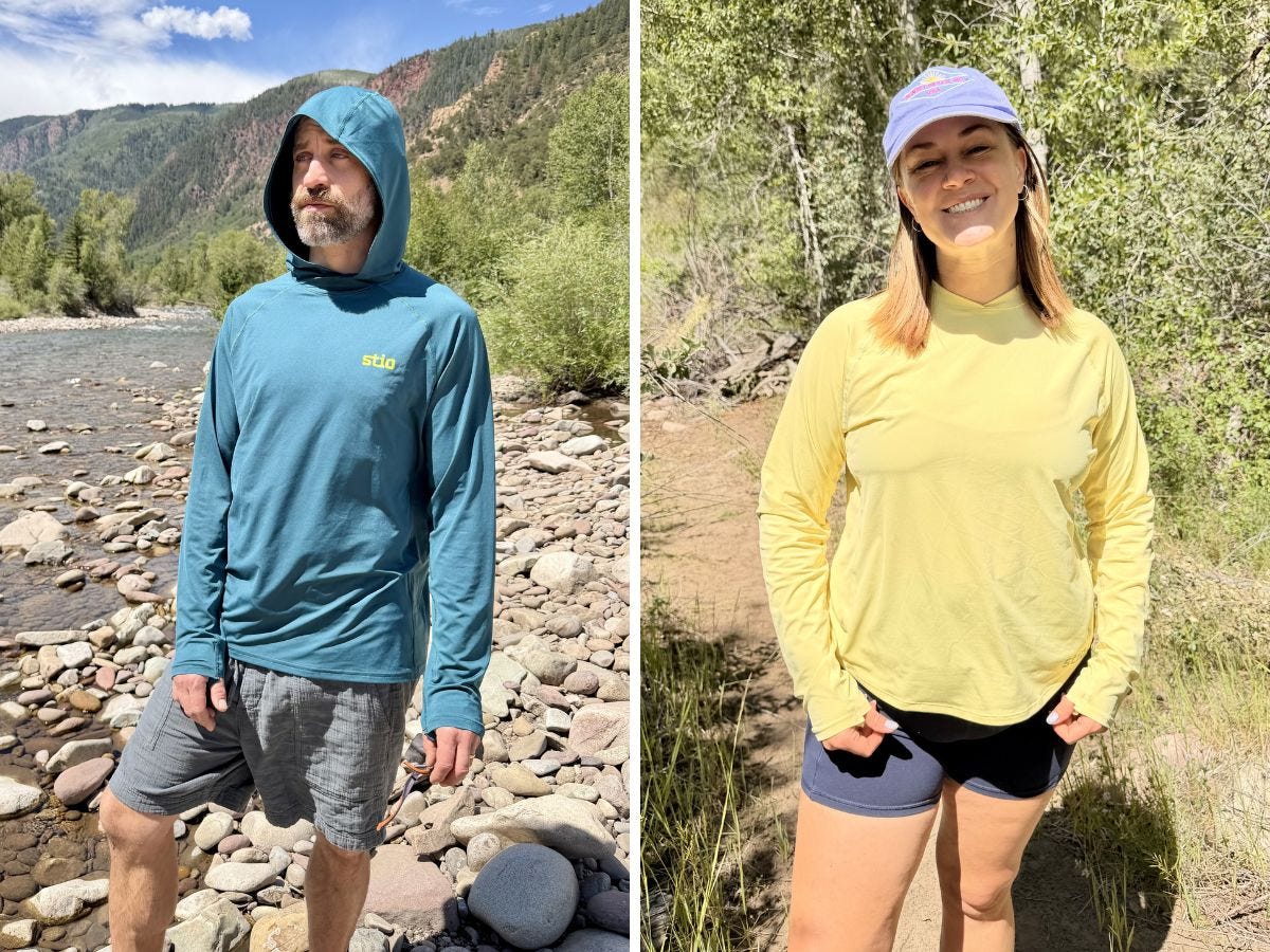 Two people wear sun hoodies while standing on rocky riverside and wooded trail in mountainous terrain.