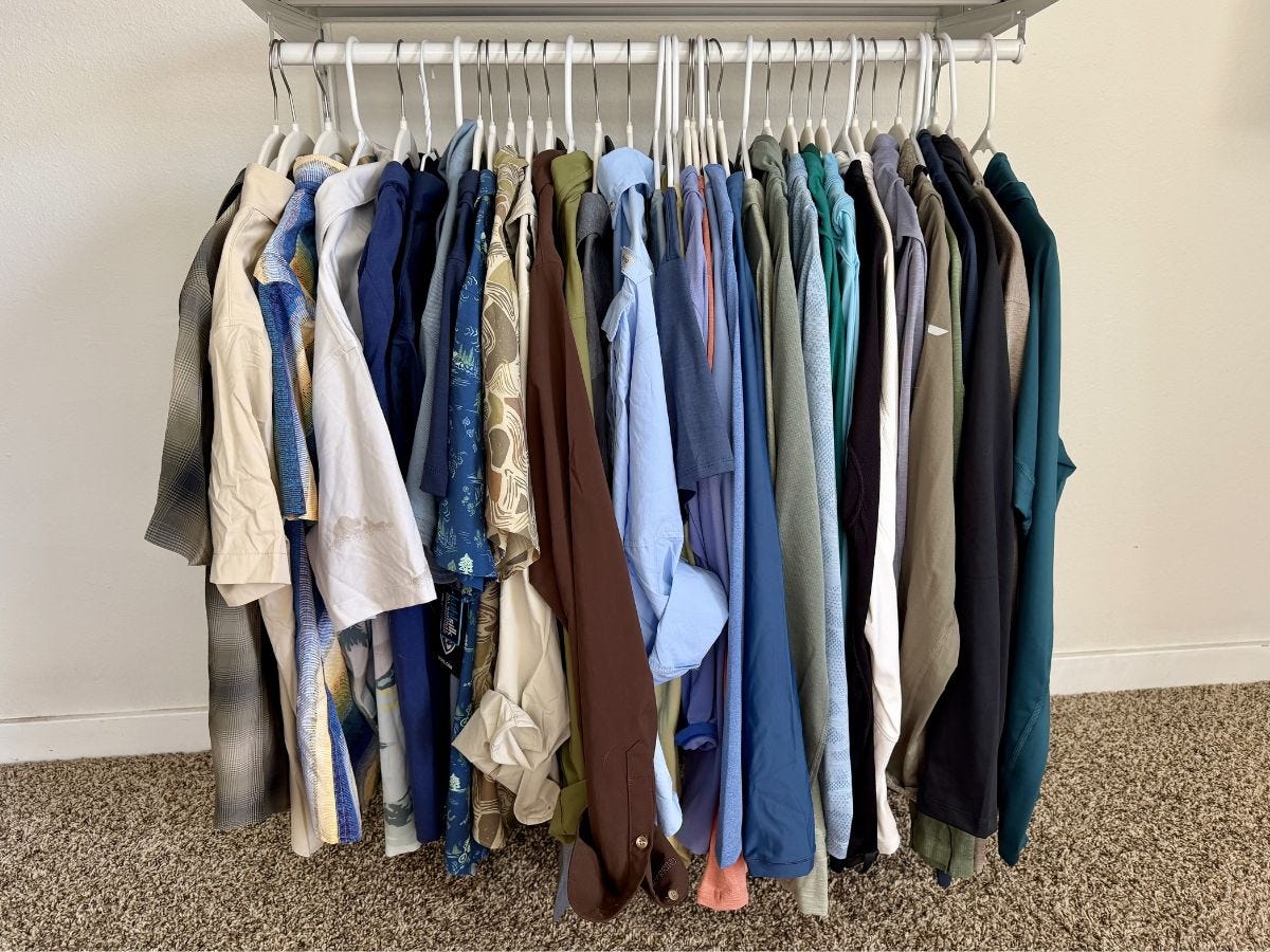 Assorted shirts and jackets hang on a freestanding clothes rack against a plain wall on carpet.
