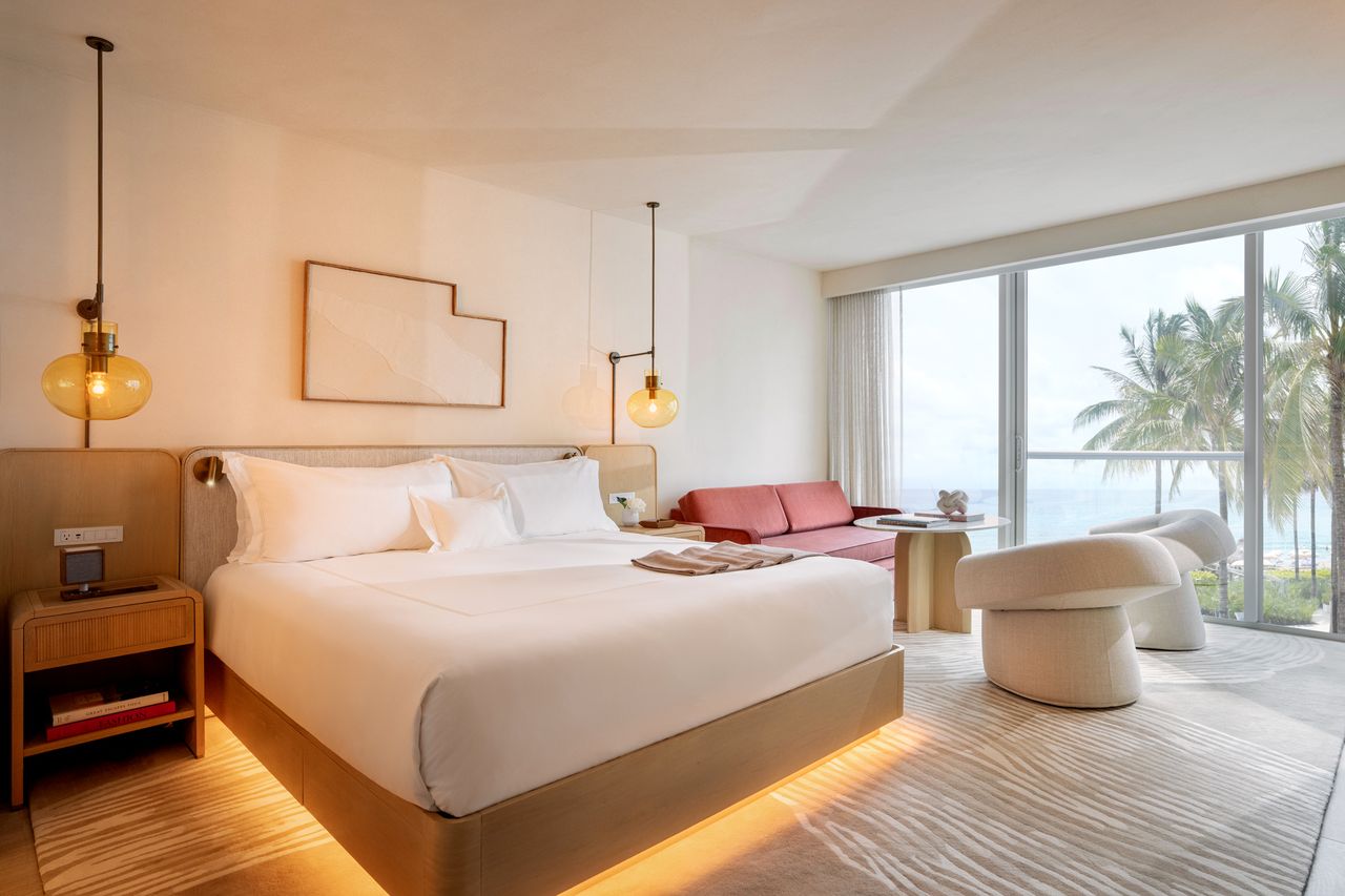A bright hotel room with a bed seating area and a view of palm trees through large windows