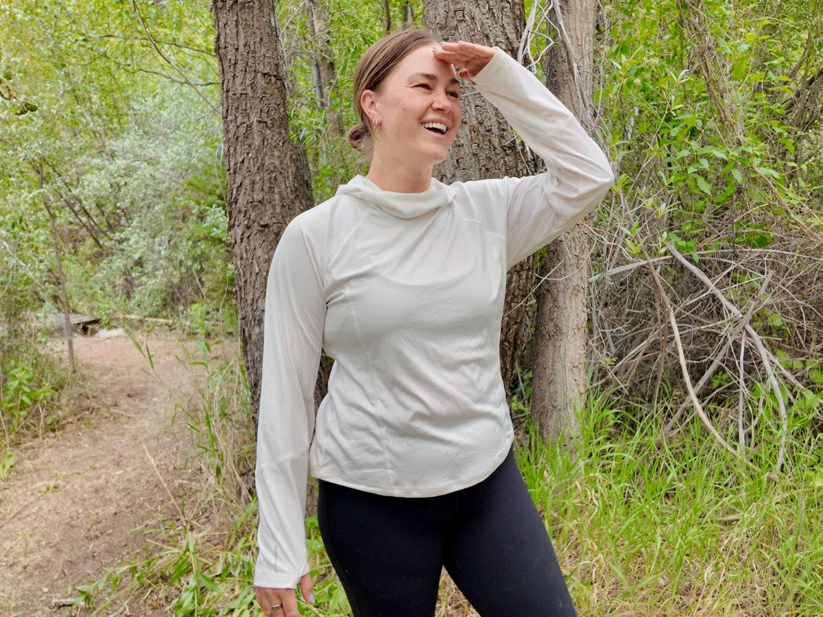 Smiling hiker shades their eyes while standing on a wooded trail among trees and green brush.