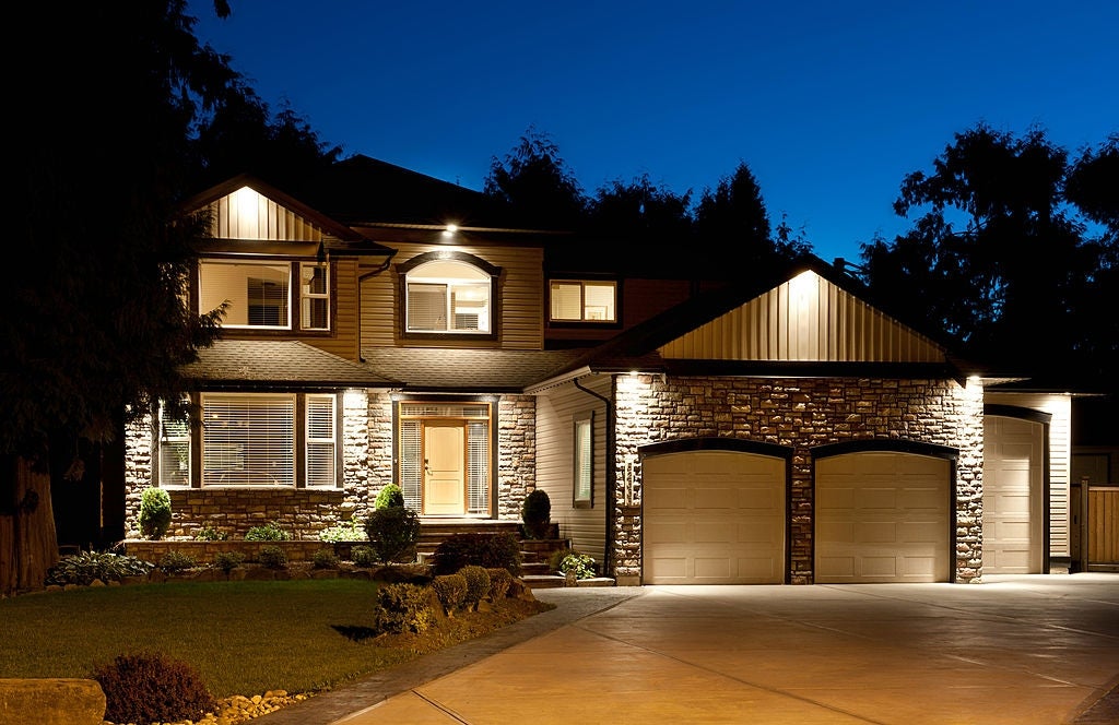 A well-lit, two-story suburban house with a stone facade and two-car garage at night, surrounded by a landscaped garden