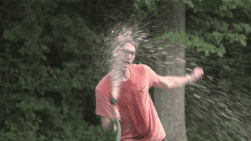 Person joyfully spraying water from hose, smiling broadly in a forest setting