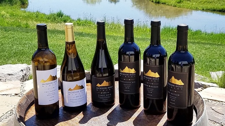 Six Jackson Hole Winery bottles of wine on a wooden barrel on stone pavers in the foreground, with green grass leading down to Spring Creek in the background.