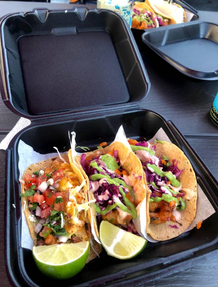 Three tacos in a takeout box with mixed toppings, including salsa, cabbage, and avocado sauce