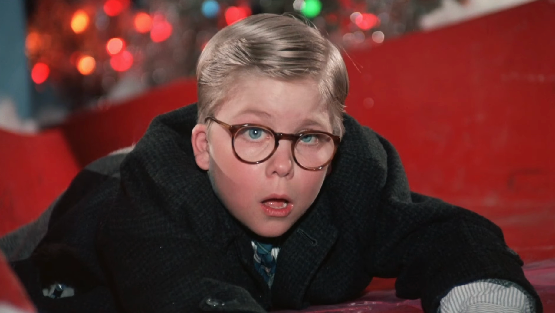 Child wearing glasses, surprised expression, lies on the ground in a holiday setting with festive lights in the background