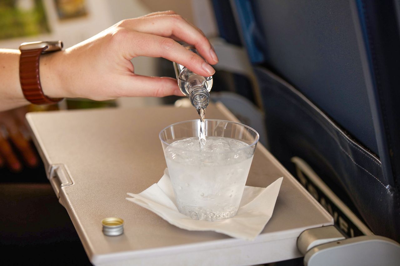 A hand pouring liquid from a small bottle into a plastic cup on an airplane tray table
