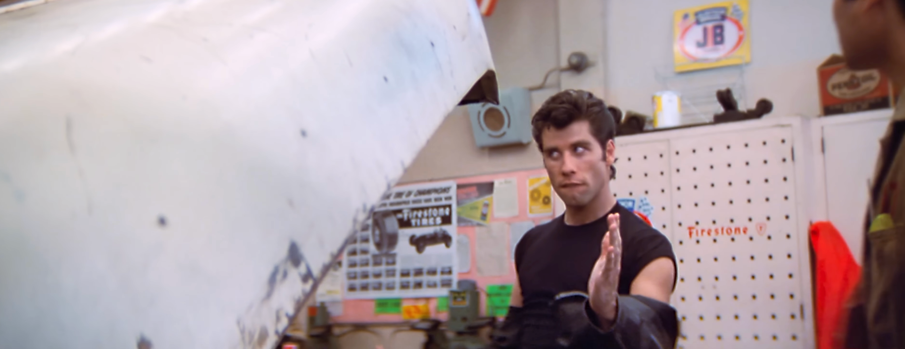 Person in a garage, gesturing toward a car hood, appearing to discuss or explain something to another person nearby