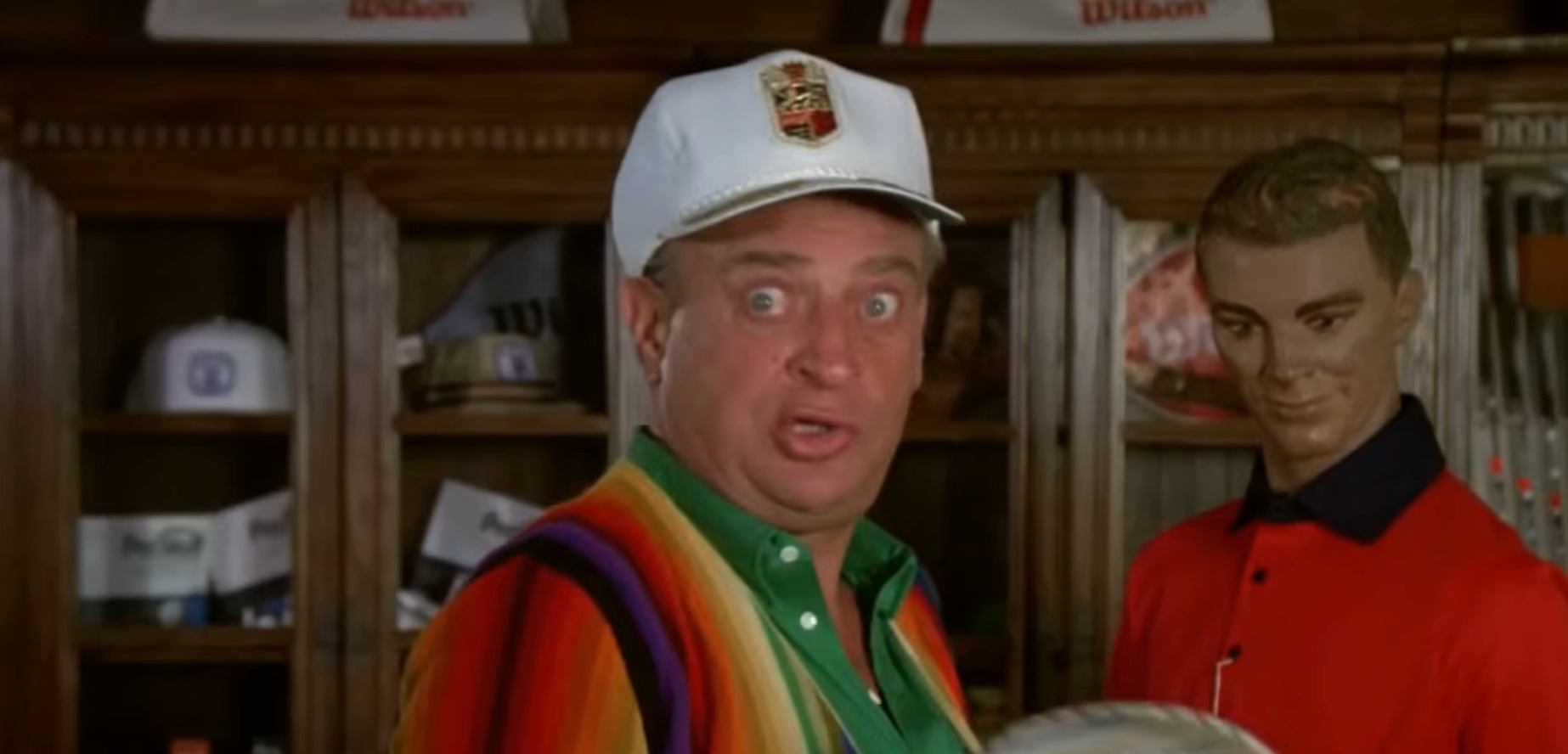 A surprised man in a colorful striped shirt and white cap stands next to a mannequin in a shop setting