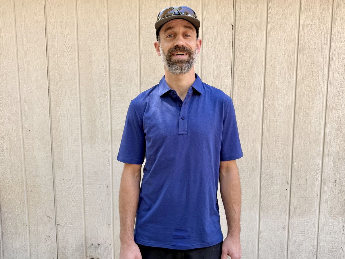 Person in a blue polo shirt and cap stands in front of a light wood-paneled wall.