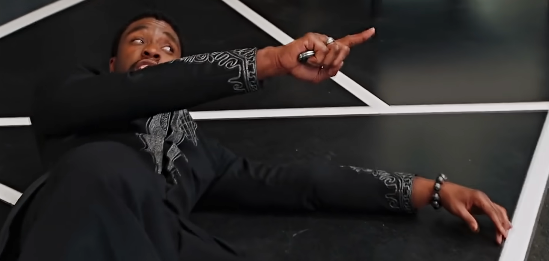 Person playfully lying on a geometric-patterned floor, pointing upwards with a smile, wearing a suit with intricate embroidery