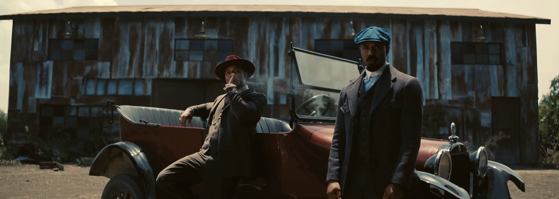 Two men in vintage attire stand by an old car in front of a weathered barn, exuding a classic, rugged style