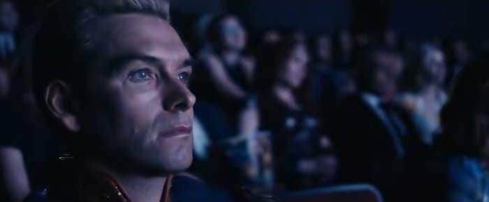 A person in a cinematic audience setting, wearing a structured jacket, intently watches something off-screen. Others are seated nearby