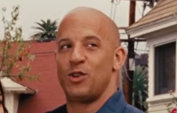 A person is outdoors, speaking with a surprised or amused expression. Background shows parts of buildings and trees