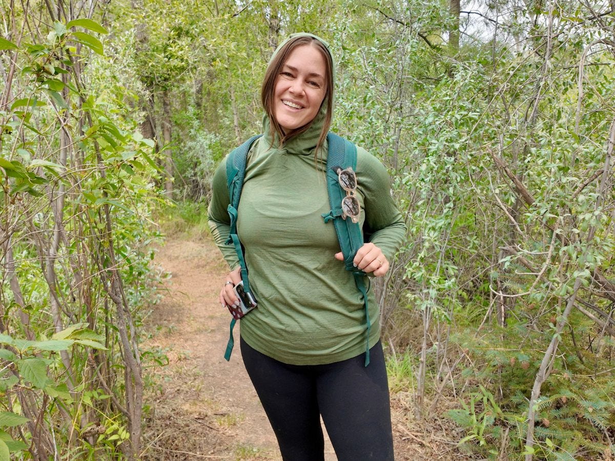 Smiling hiker wearing a green top and backpack stands on a narrow wooded trail.