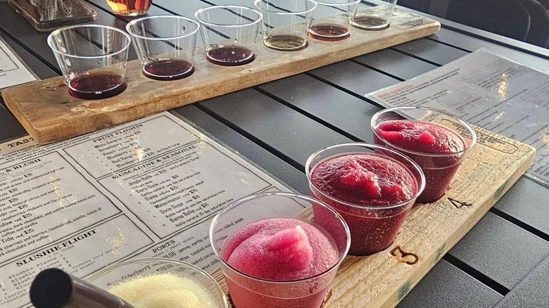 A bright-colored wine slushie flight in the foreground with a wine flight in the background, both served in clear cups on wooden boards, on a table with wine menus.