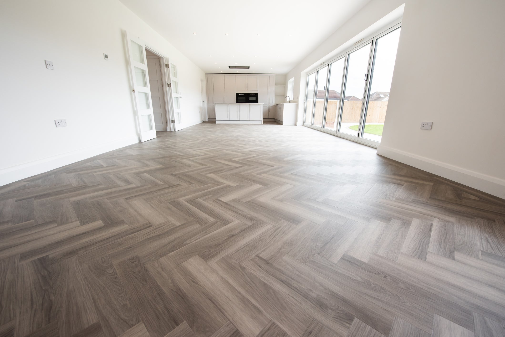 Spacious, unfurnished room with herringbone-pattern wood flooring, large windows, and a modern kitchen island in the background