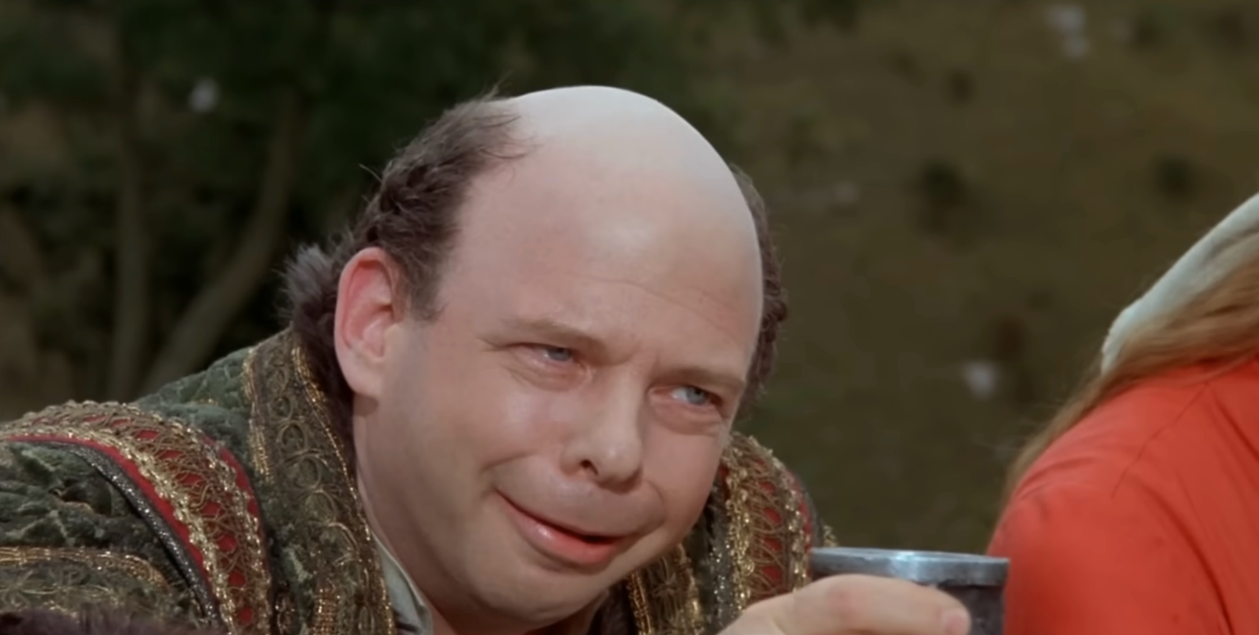 A person with a bald head smiles mischievously while holding a cup, in a scene from a movie