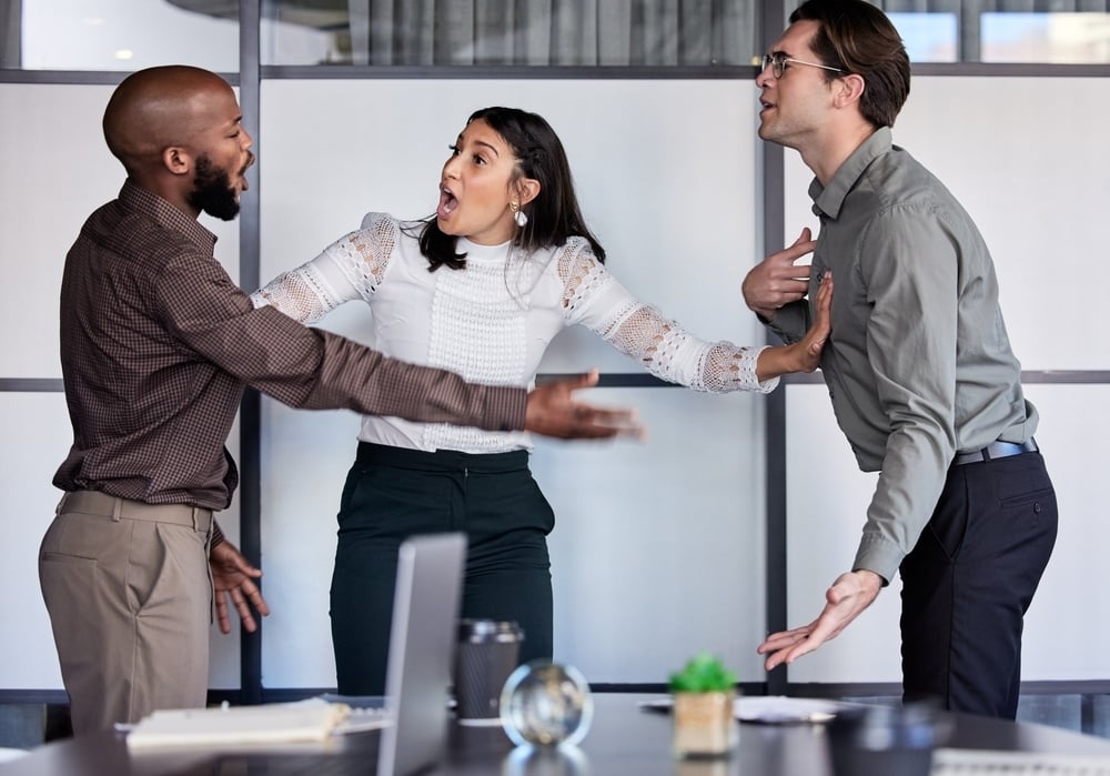 A woman stands between two men, all yelling and gesturing during a tense office argument.
