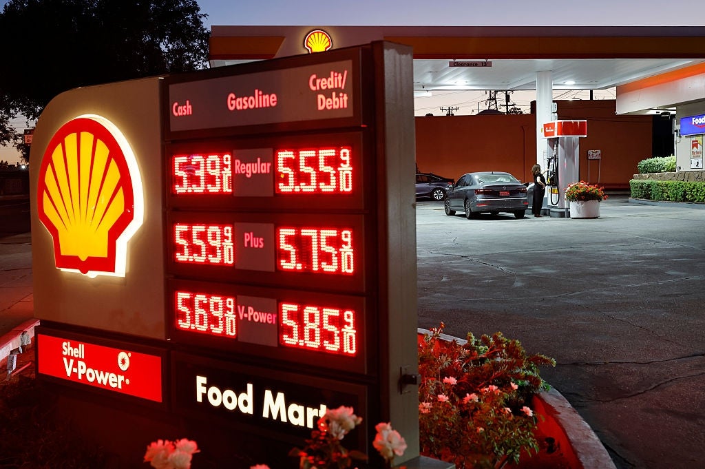 A gas station sign displays fuel prices: Regular $5.55, Plus $5.75, V-Power $5.85. A car is parked by the fuel pumps at the station