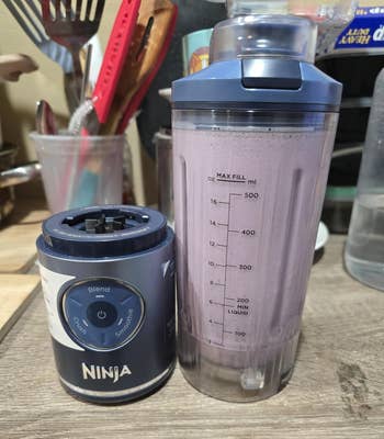 A blender base next to a smoothie-filled cup. Measuring lines show liquid levels. Kitchen utensils and items are in the background