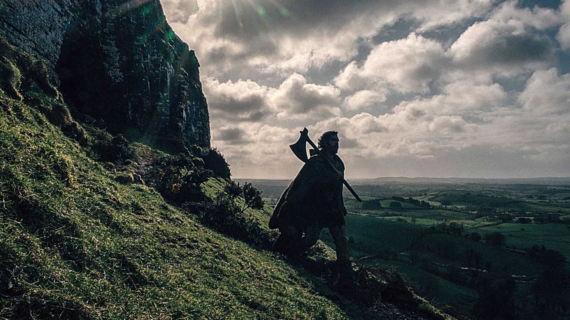 Gawain (Dev Patel) walking down a hill, carrying an axe in The Green Knight.