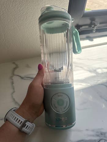 Person holding a portable Ninja blender with blending modes displayed, suitable for smoothie making, placed on a marble countertop near a window