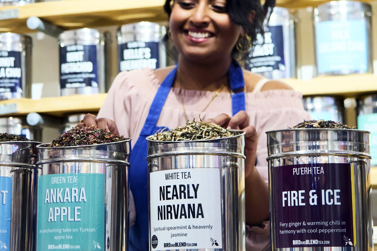 Smiling person arranging tea tins labeled Ankara Apple Nearly Nirvana and Fire Ice on a display