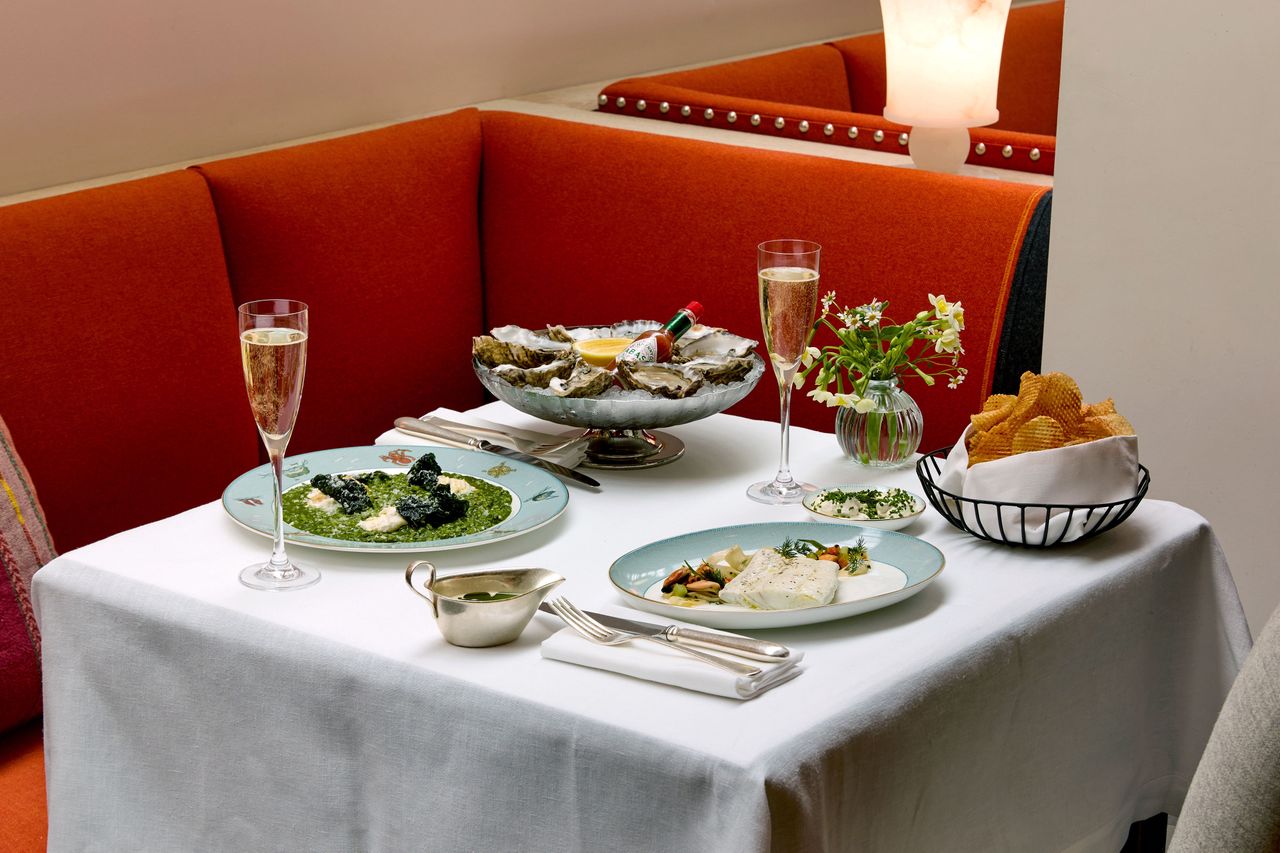 A dining table with plates of oysters spinach fish dishes glasses of champagne and a basket of bread in a restaurant seating area