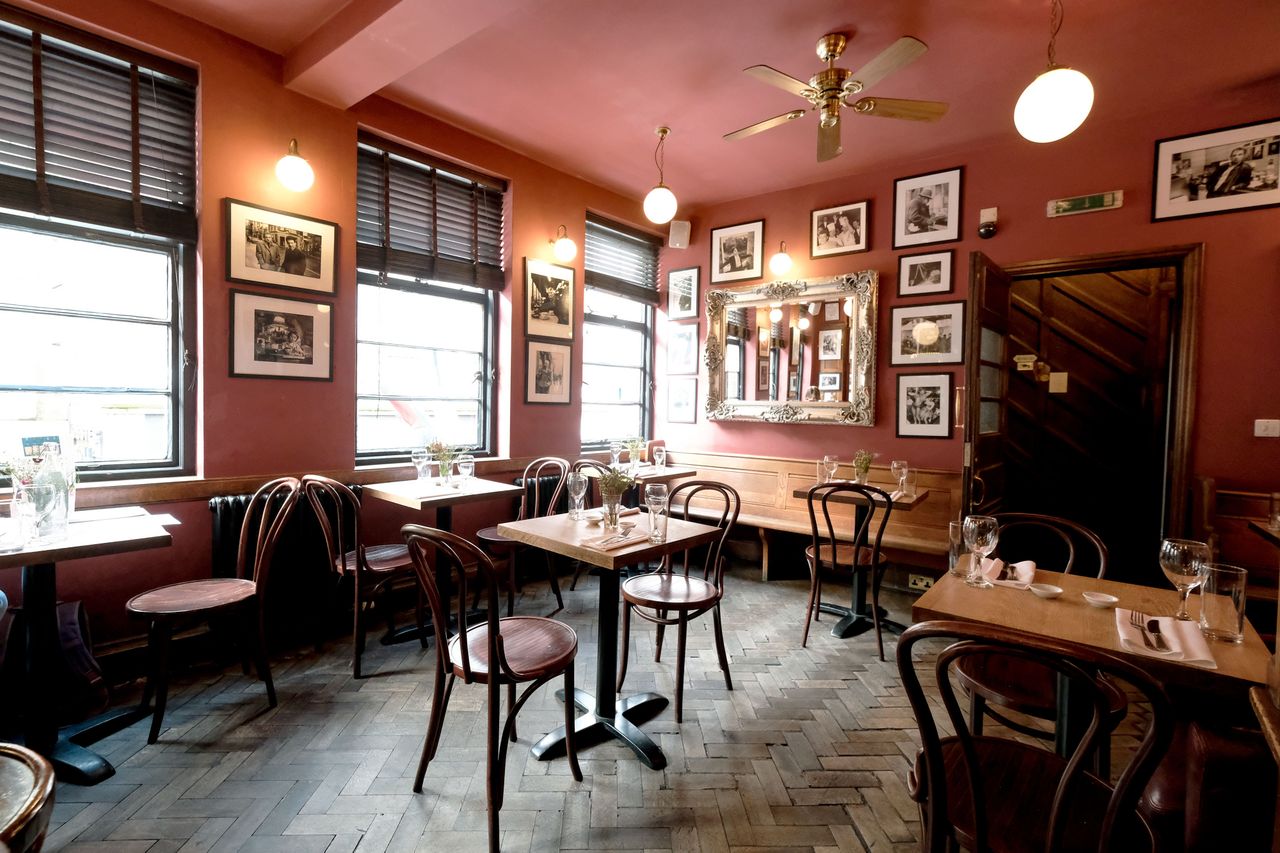 A cozy restaurant interior with wooden chairs tables set for dining framed pictures on the walls and a mirror in the room