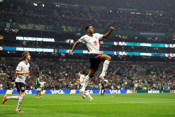 Soccer player in white jersey jumps in celebration during a match. Teammates and spectators in stadium visible in the background