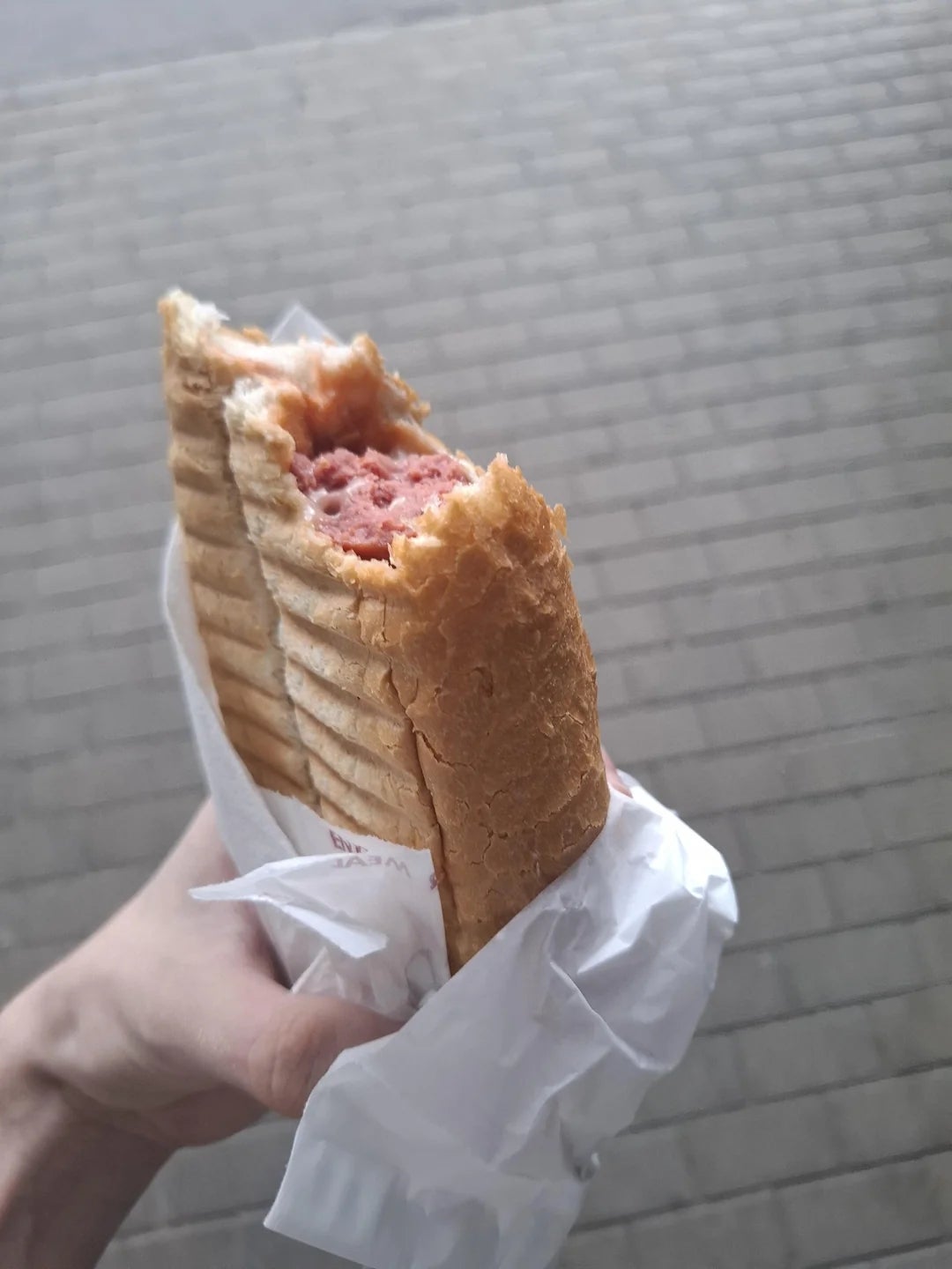 A hand holds a partially eaten sandwich with grilled bread and a visible meat filling, wrapped in white paper