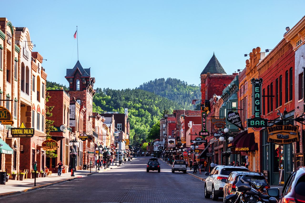 A street in a town with cars parked along buildings and a hill in the background
