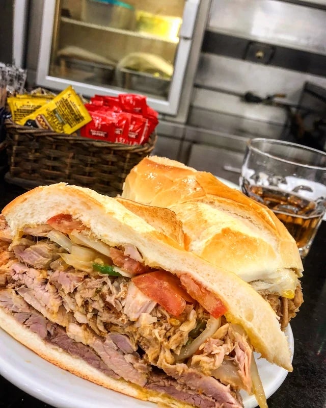 Sandwich filled with shredded meat, onions, and tomatoes on a plate beside a drink and condiments basket in a restaurant setting