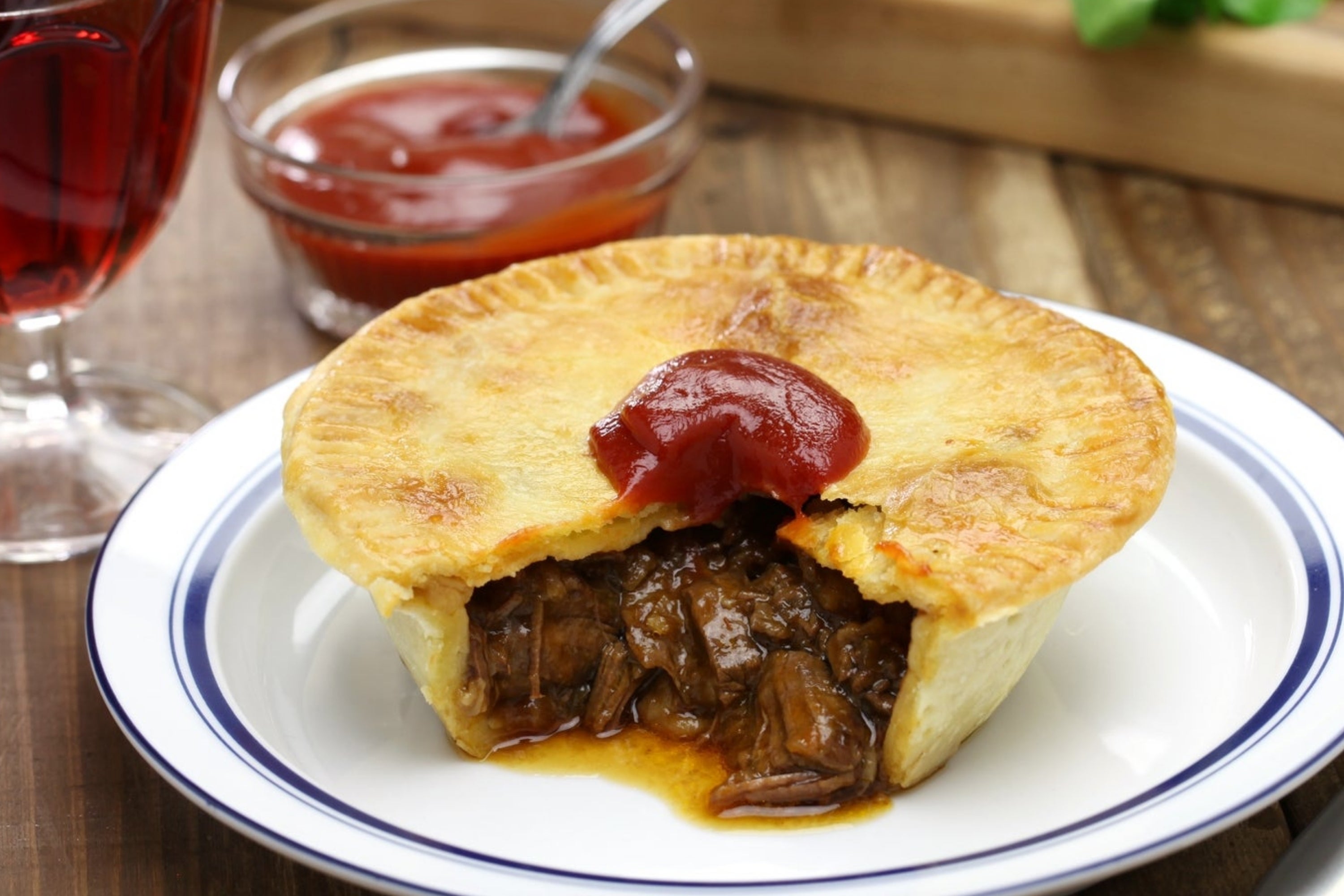 Meat pie on a plate with a bite taken out, topped with ketchup. Glass of red wine, utensils, and garnish on a wooden table