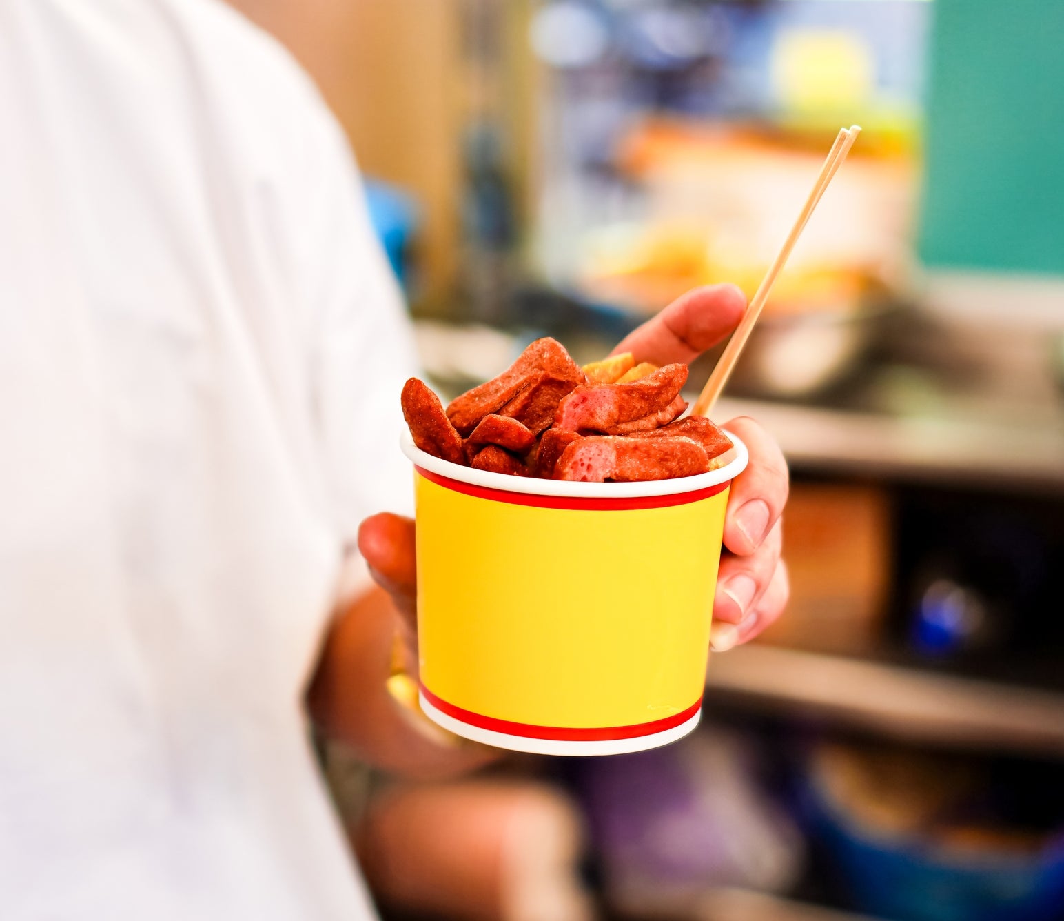 Person holds a cup of sliced pepperoni with a wooden stick, likely from a food stand or market
