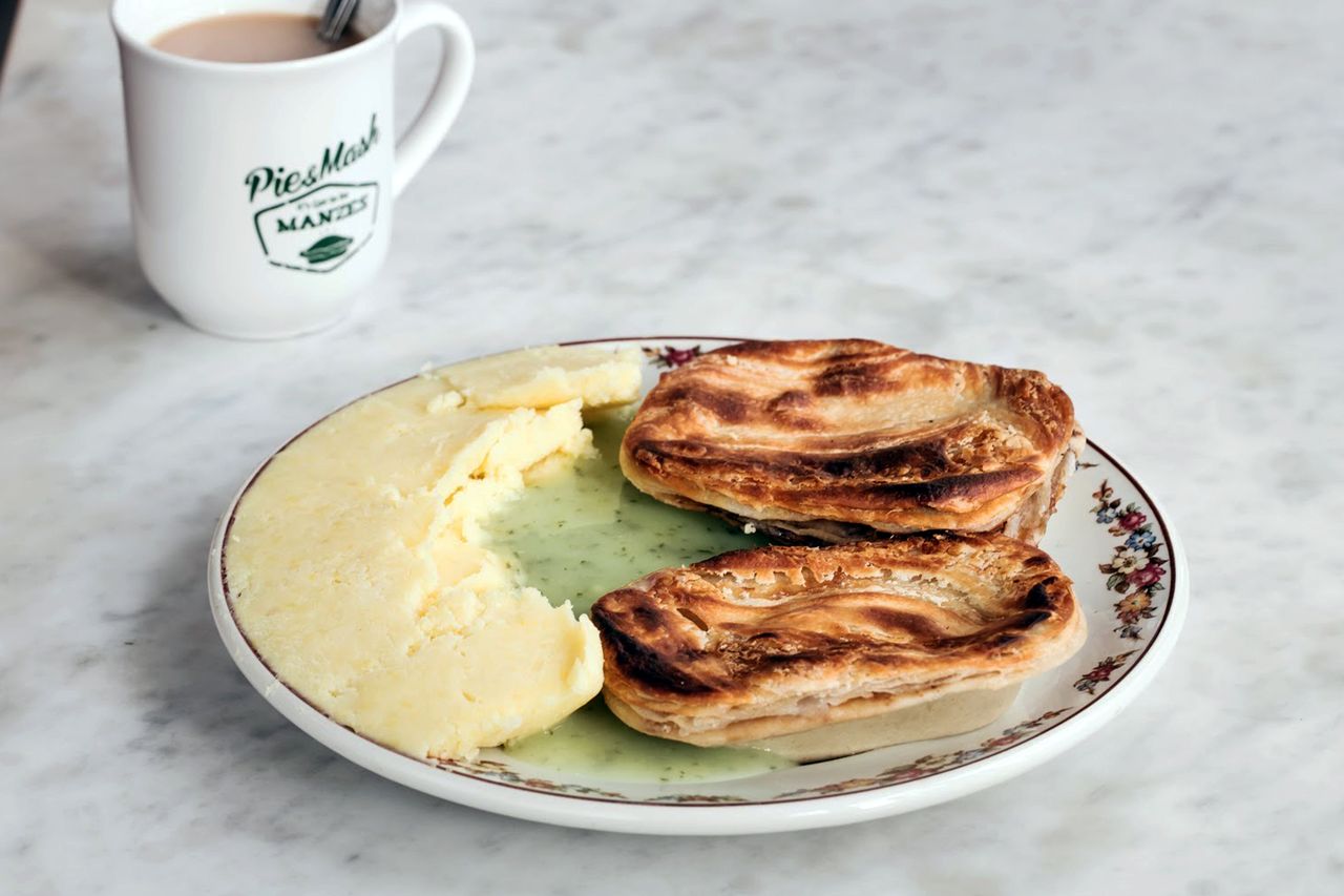 A plate with two pies mashed potatoes and a green sauce next to a mug labeled Pie Mash on a marble surface