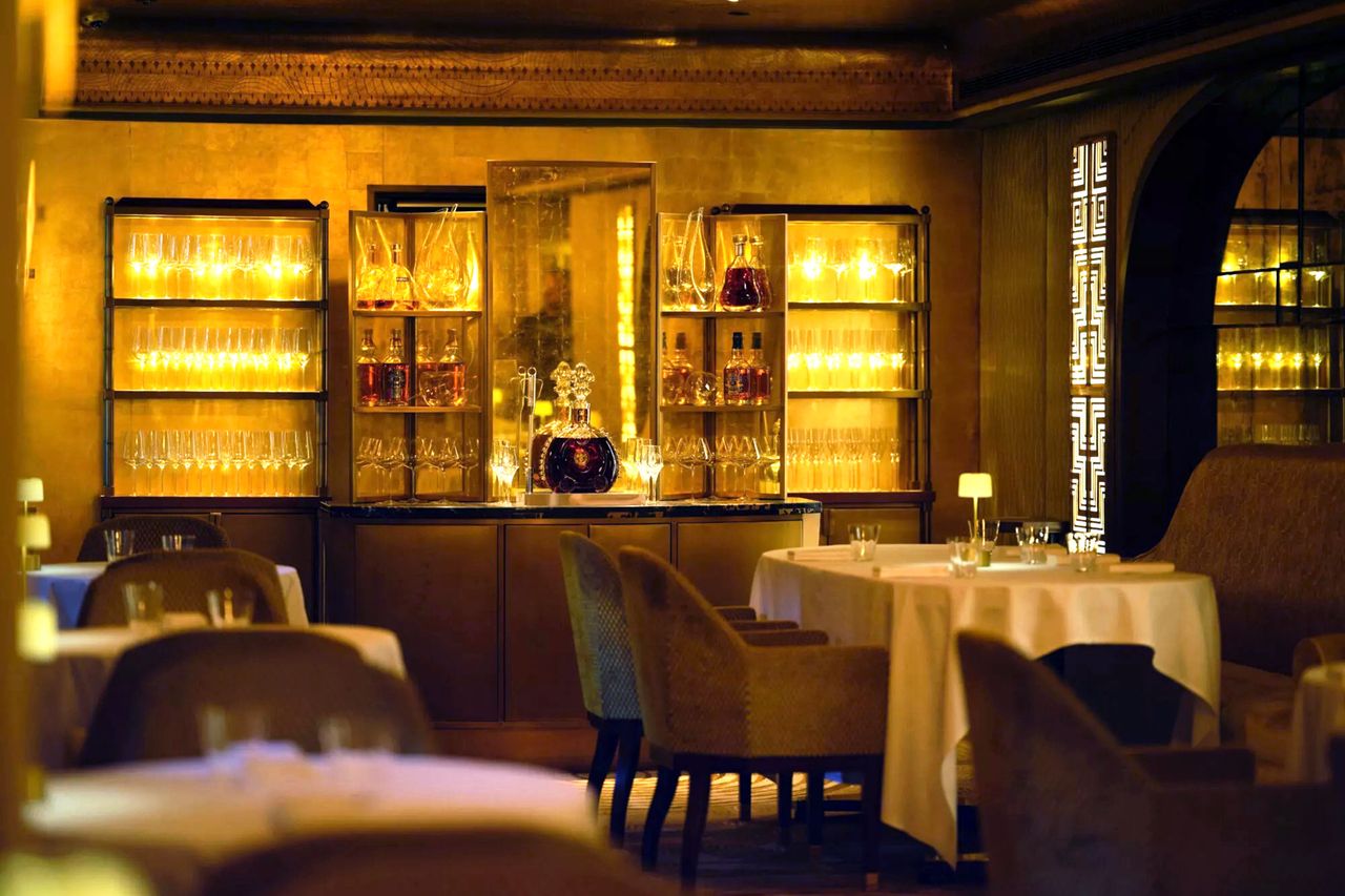 Elegant interior of Restaurant 1890 with round tables and upholstered chairs shelves of glassware and decorative bottles illuminated in the background