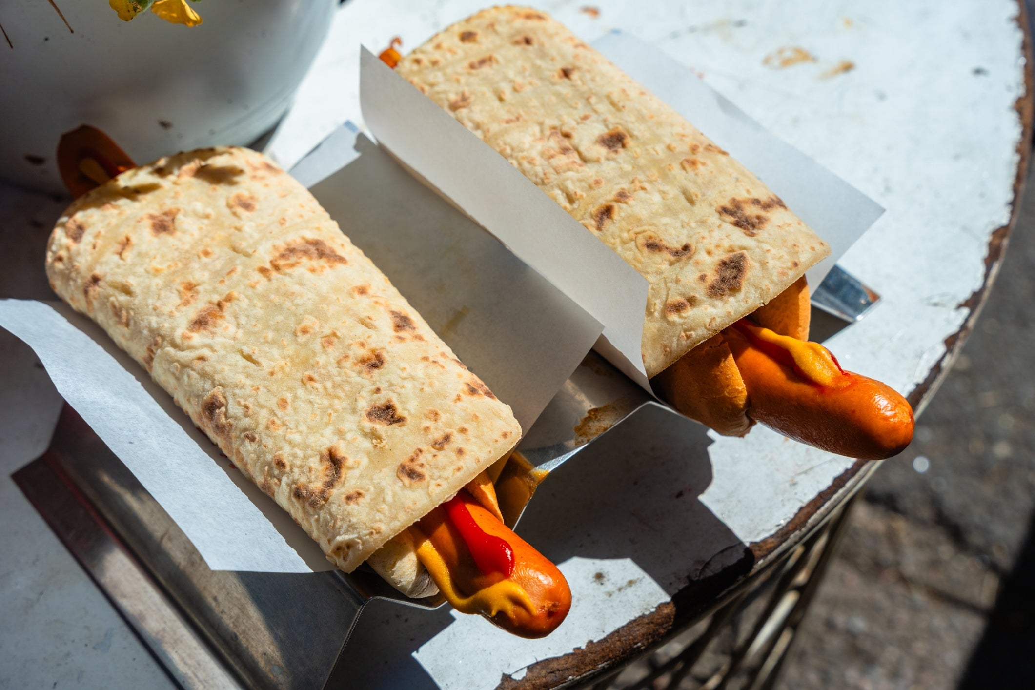 Two hot dogs wrapped in flatbread with mustard, placed in paper trays on a round table