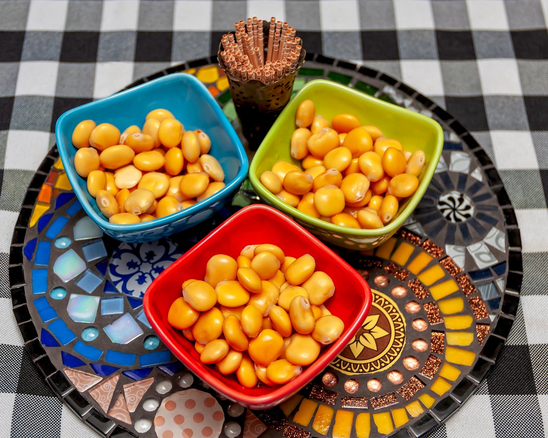 Three colorful bowls of lupin beans on a mosaic-patterned tray, with a small cup of toothpicks on a checkered tablecloth