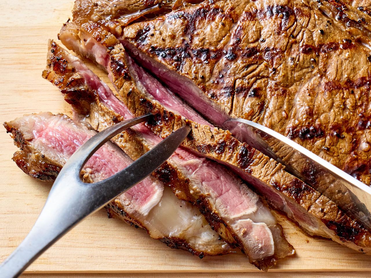 A grilled steak being sliced on a wooden cutting board with a carving fork and knife