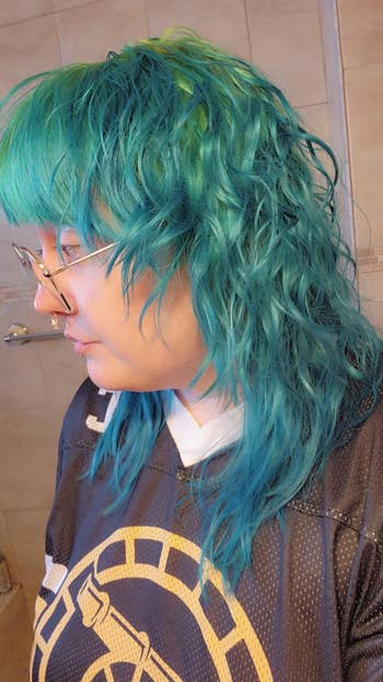 Person with wavy teal hair wearing glasses and a sports jersey with a circular emblem, standing indoors
