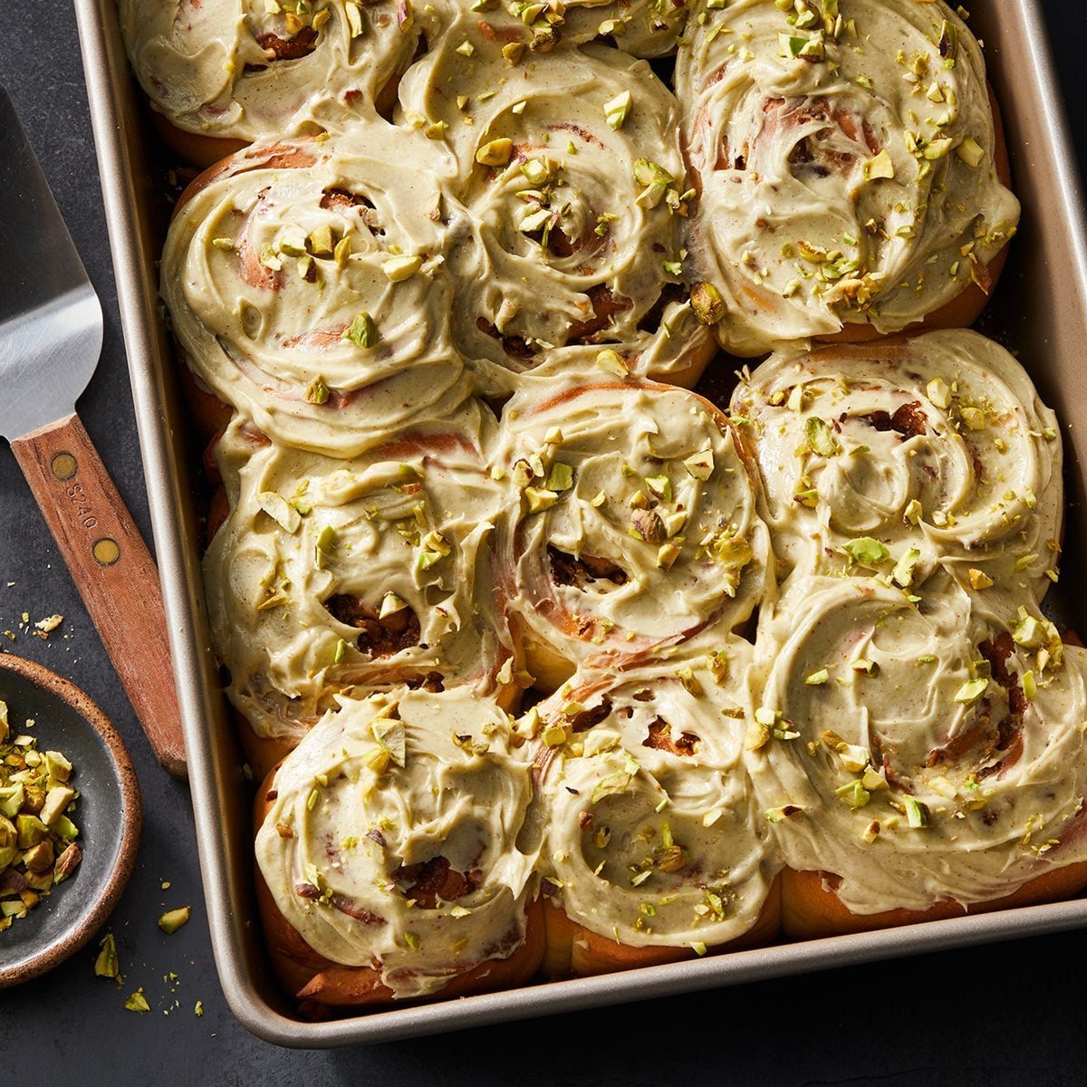 A baking tray filled with swirled pistachio frosted cinnamon rolls, topped with chopped pistachios. A serving spatula and a small bowl of extra chopped pistachios sit nearby on a dark surface.