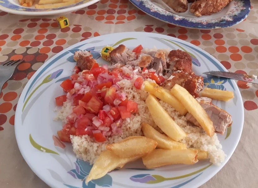 Plate of couscous with diced tomatoes, onions, fries, and grilled meat on a table with food and utensils