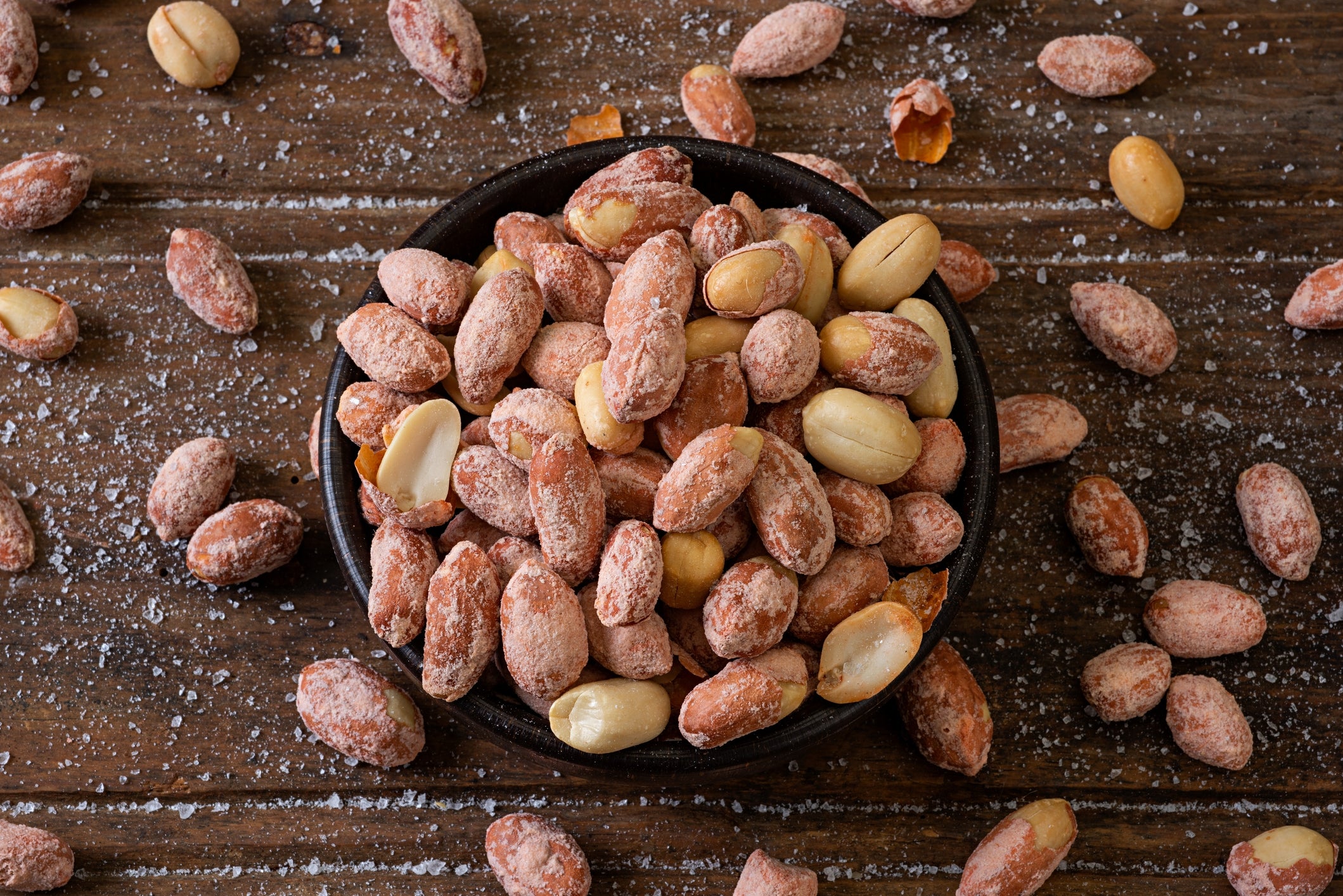 Bowl filled with salted peanuts, some scattered on a wooden surface